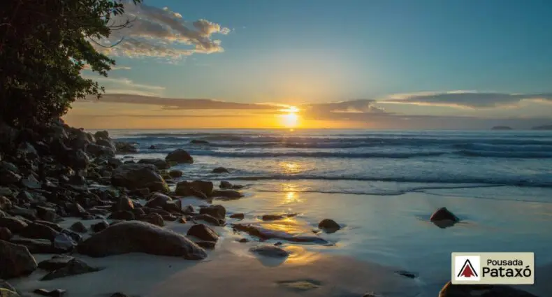 Praia das Toninhas ao pôr do sol, com a luz do sol refletindo na areia molhada e nas ondas, com o logotipo da "Pousada Pataxó" no canto, para representar pousadas na Praia das Toninhas em Ubatuba.