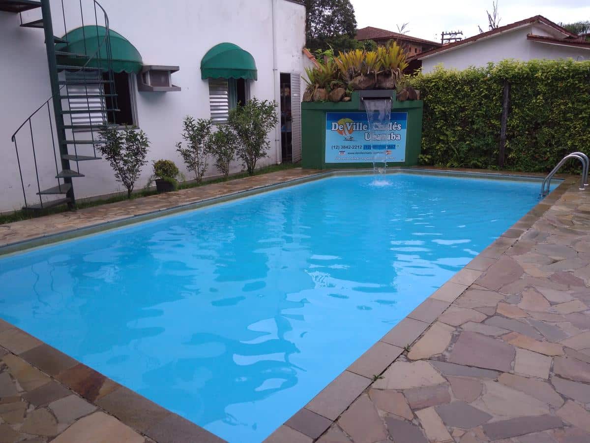 Piscina externa retangular do De Ville Chalés com água limpa, deck de pedra, plantas verdes e um prédio com toldos verdes nas proximidades.