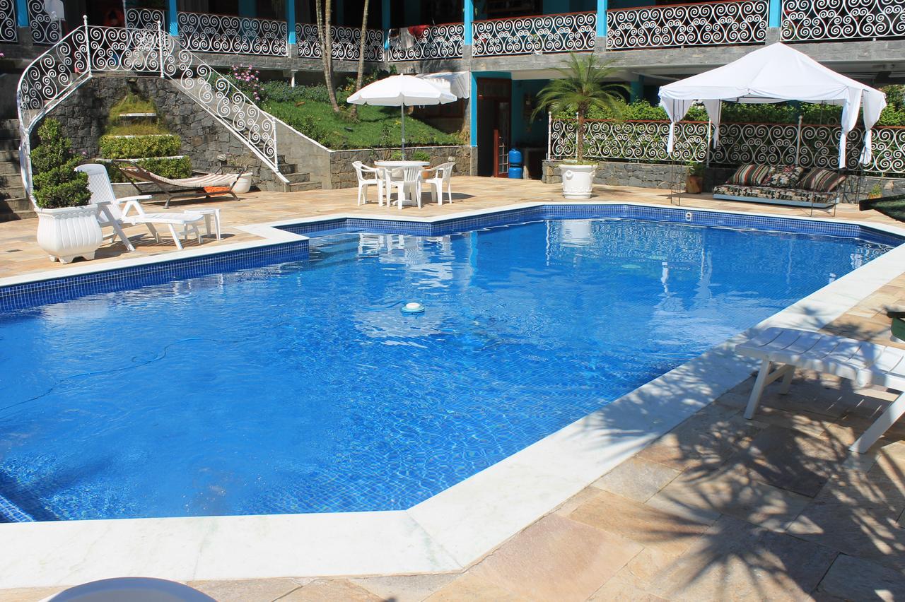 Piscina externa do Espaço Blue House com água azul clara, cercada por cadeiras, mesas, guarda-sóis e grades decorativas no pátio.