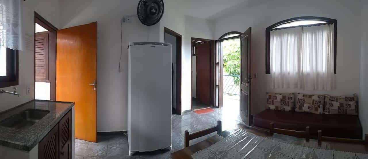 Pequena cozinha e sala de estar do Excellence Apart com geladeira, pia, sofá marrom, portas de madeira, janela e piso de ladrilho.