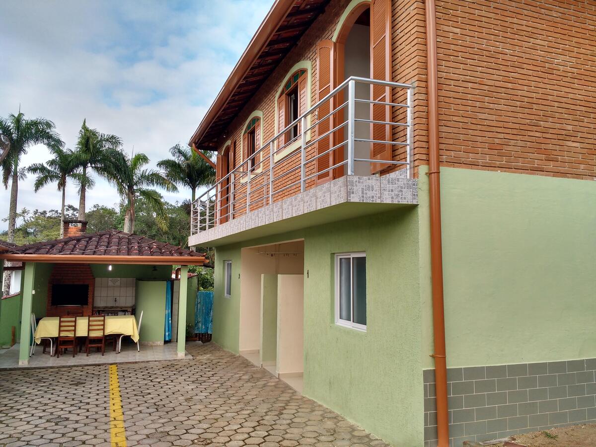 Uma casa de dois andares com varanda e paredes inferiores verdes, ao lado de um pátio coberto com mesa e cadeiras. Esse é o Praialar Apartamentos Ubatuba.