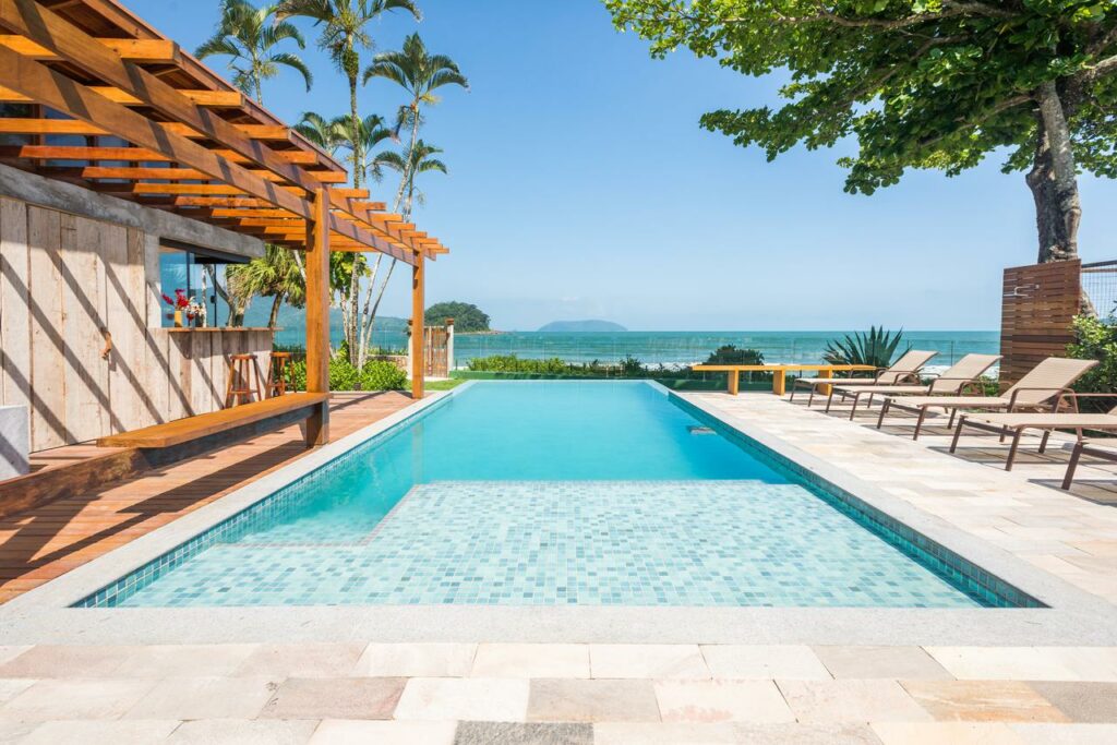Piscina da Villa Sapê Pousada, uma piscina ampla com vista para a praia, com espreguiçadeiras e bancos dos dois lados, além das árvores