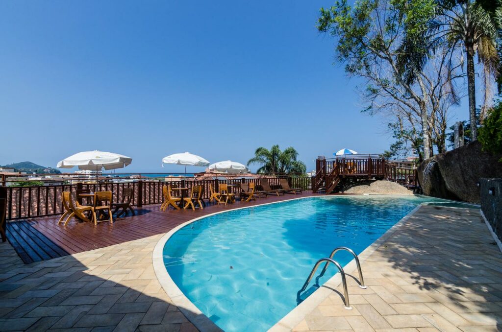 Piscina com deck, mesas, cadeiras e guarda-sóis no Hotel Coquille, um dos hotéis em Ubatuba, sendo possível avistar o mar bem ao fundo
