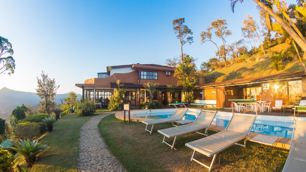 Pousada Le Siramat com casa ao fundo, com piscina e vegetação ao entorno, com cadeiras de praia em primeiro plano em um dia azul para representar as pousadas em Petrópolis.