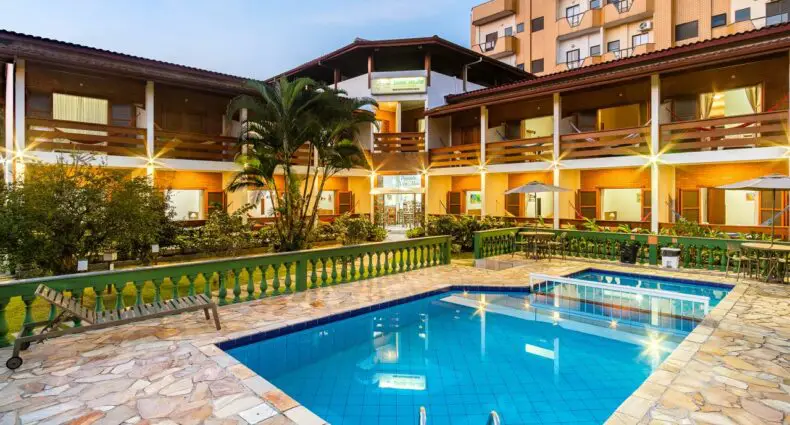 Uma piscina externa retangular com degraus, cercada por um pátio de pedra e um prédio de dois andares do hotel ao anoitecer.