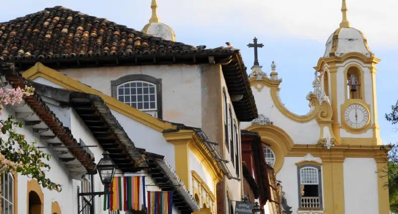 Prédios coloniais com telhados de telha em uma rua estreita; uma igreja amarela e branca com uma torre de relógio ao fundo, em Tiradentes, para representar aibnb em Tiradentes