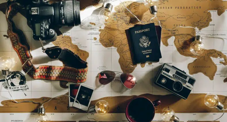 Mapa mundi com uma câmera, um óculos de sol, passaporte, café e fotos em cima para representar o seguro saúde viagem. - Foto: Charlotte Noelle via Unsplash