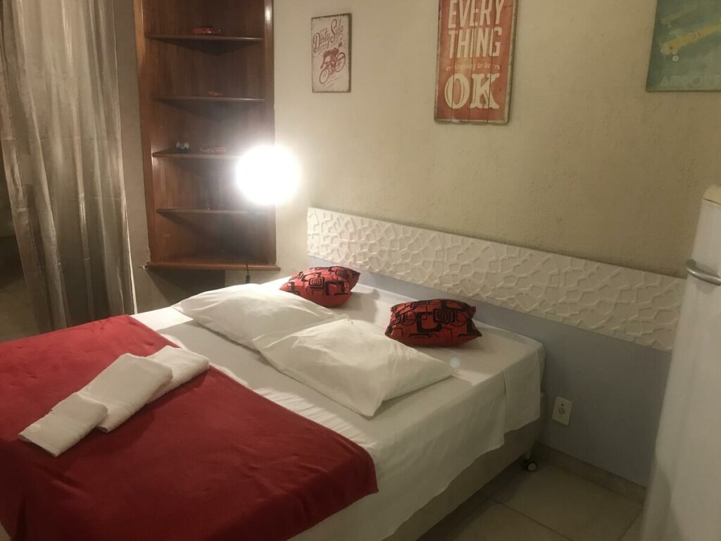 Quarto da Albergo Alegria, com uma cama de casal com jogo de cama vermelho e branco e uma prateleira de canto no lado esquerdo com um abajur aceso na frente