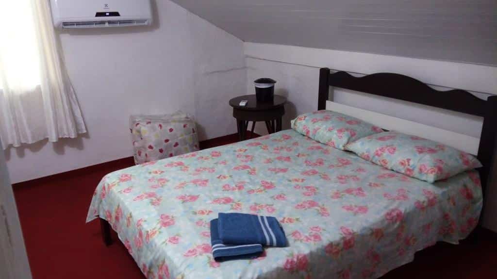 Quarto duplo da Austria Hostel & Pousada, de 14 m², com uma cama de casal, duas toalhas dobradas em cima, uma mesinha redonda ao lado, um edredom dobrado dentro de um pacote de plástico transparente ao lado e um ar-condicionado na parede esquerda