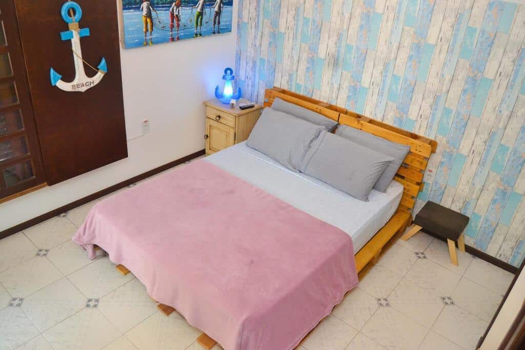 Quarto da CanCoq Guest House, com uma cama de casal rústica feita de palet, ao lado há um banquinho baixo e no outro lado da cama tem uma escrivaninha de madeira clara com uma luminária azul acesa no formato de uma âncora para ilustrar as pousadas em Balneário Camboriú