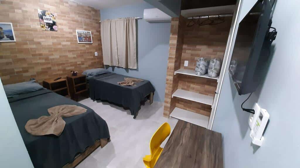 Interior do quarto triplo da Casa da Maga, de 16 m², com uma cama de casal e uma de solteiro, duas escrivaninhas ao lado das camas, um TV suspensa na parede, ar-condicionado e uma mesa com uma cadeira amarela