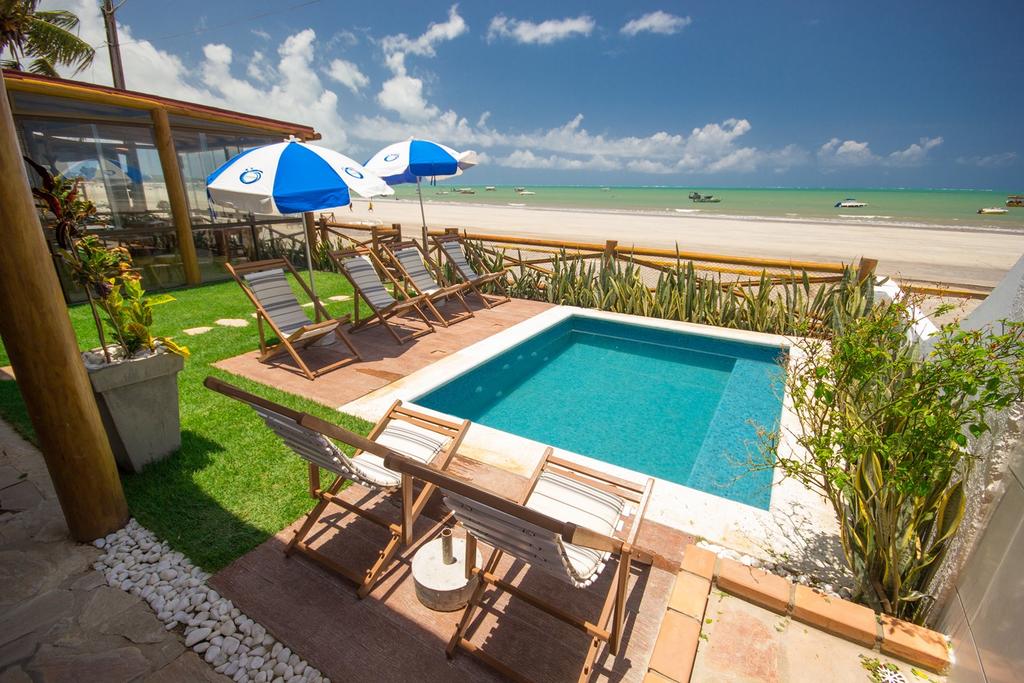 Piscina quadrada com espreguiçadeiras e guarda-sóis em volta, localizados à beira-mar com areia branca e mar verde cristalino para ilustrar as pousadas em Alagoas