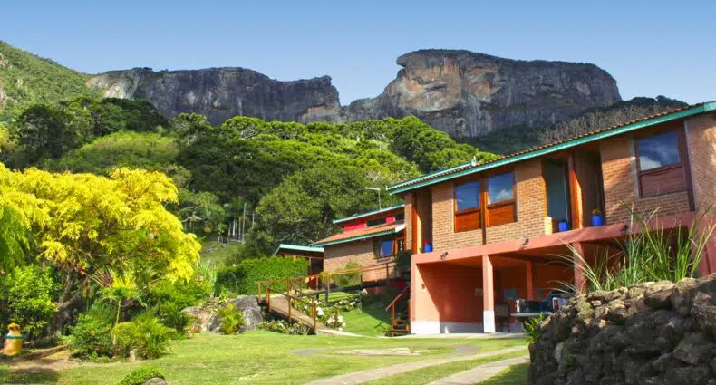 Fachada da Hospedaria Estação Píccola em dois andares no meio de um grande gramado verde com árvores verdes ao redor e Vista da Pedra do Baú em uma das pousadas em São Bento do Sapucaí.
