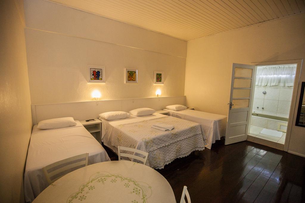 Quarto do Hotel Pousada XV, com uma cama de casal e duas de solteiro, uma mesa redonda com quatro cadeiras e uma porta que dá acesso ao banheiro com hidro