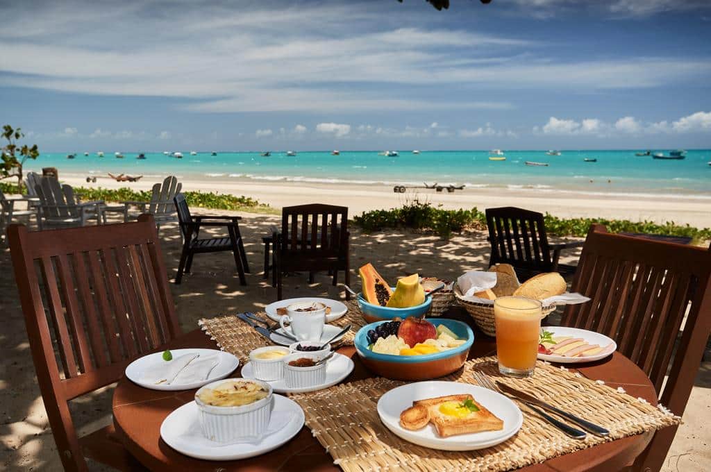 Mesa com café da manhã na areia de uma praia de água azulada com vários barcos espalhados. A areia é branca e o céu está azulado com nuvens brancas para ilustrar as pousadas em Alagoas