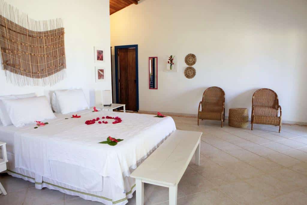 Quarto da Pousada Borapirá, com cama de casal com pétalas vermelhas em cima, duas poltronas e uma decoração rústica com peças artesanais penduradas na parede