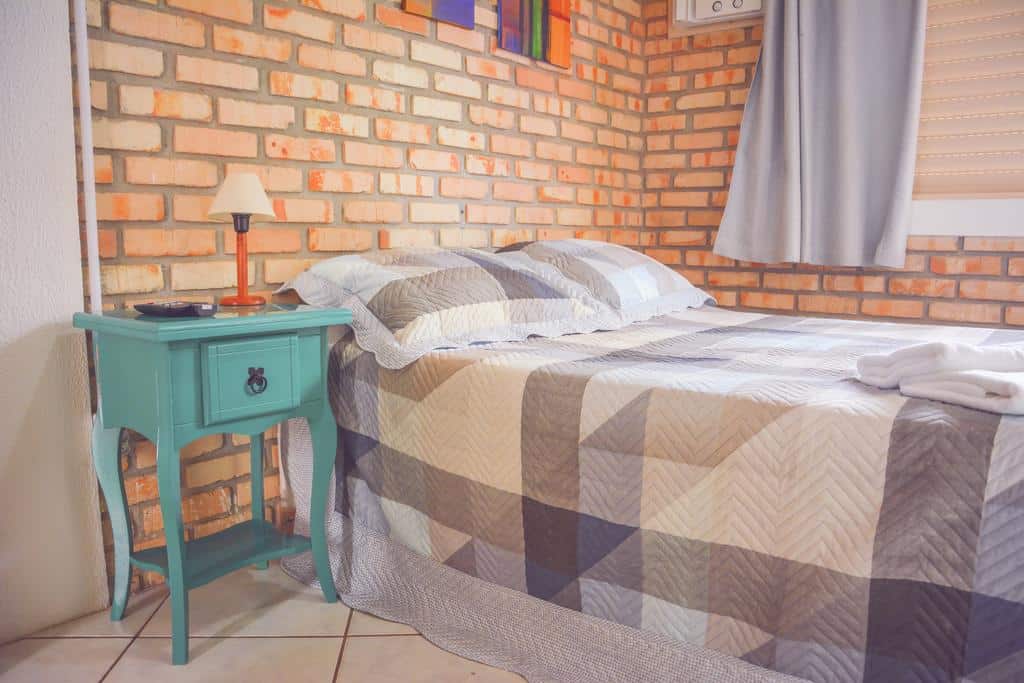 Quarto da Pousada Canto da Brava, com uma cama de casal, uma escrivaninha retrô verde ao lado com um abajur em cima e as paredes são de tijolinhos à vista