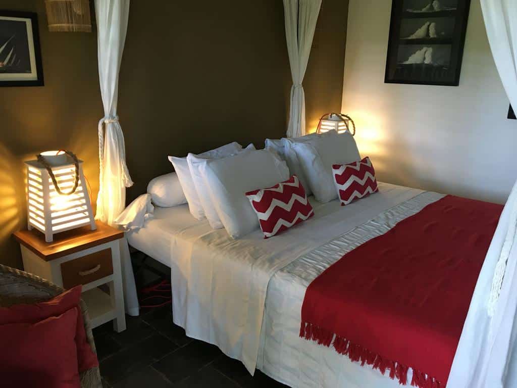 Cama de casal em uma das pousadas em São Miguel dos Milagres, a Côté Sud, com roupas de cama brancas e detalhes em vermelho, além de mesinha de cabeceira e abajures estilizados a cada lado da cama