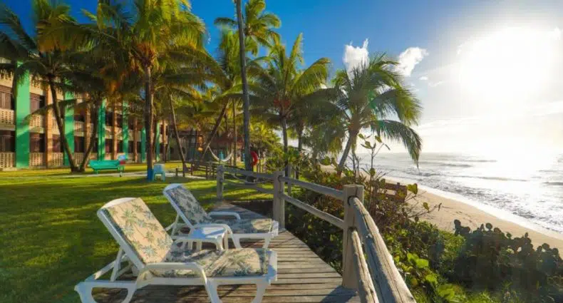 Espreguiçadeiras em um deck de madeira têm vista para uma praia de areia com palmeiras sob um céu azul claro e sol brilhante.