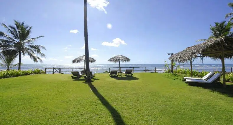 Amplo gramado com espreguiçadeiras na Pousada Farol das Tartarugas com guarda-sóis de palapa, de frente para uma praia de areia e para o oceano sob um céu azul claro. Ilustra o post de pousadas na Praia do Forte.