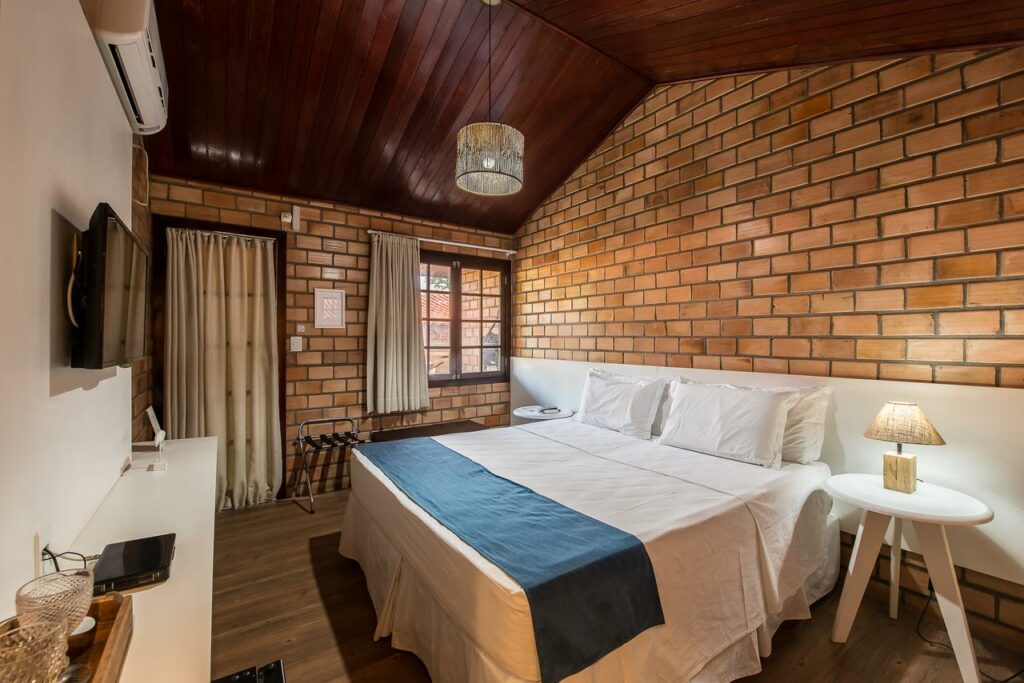 Quarto da Pousada Filó, com parede de tijolinhos, cama de casal ampla, mesa de cabeceira com abajur, quatro travesseiros, lençóis brancos com peseira azul