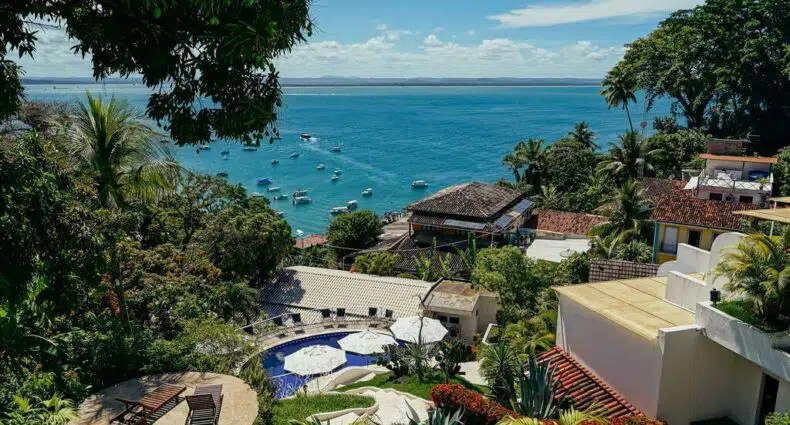 Vista do topo de uma colina com vista para uma piscina, vegetação exuberante e barcos em um oceano azul sob um céu claro.