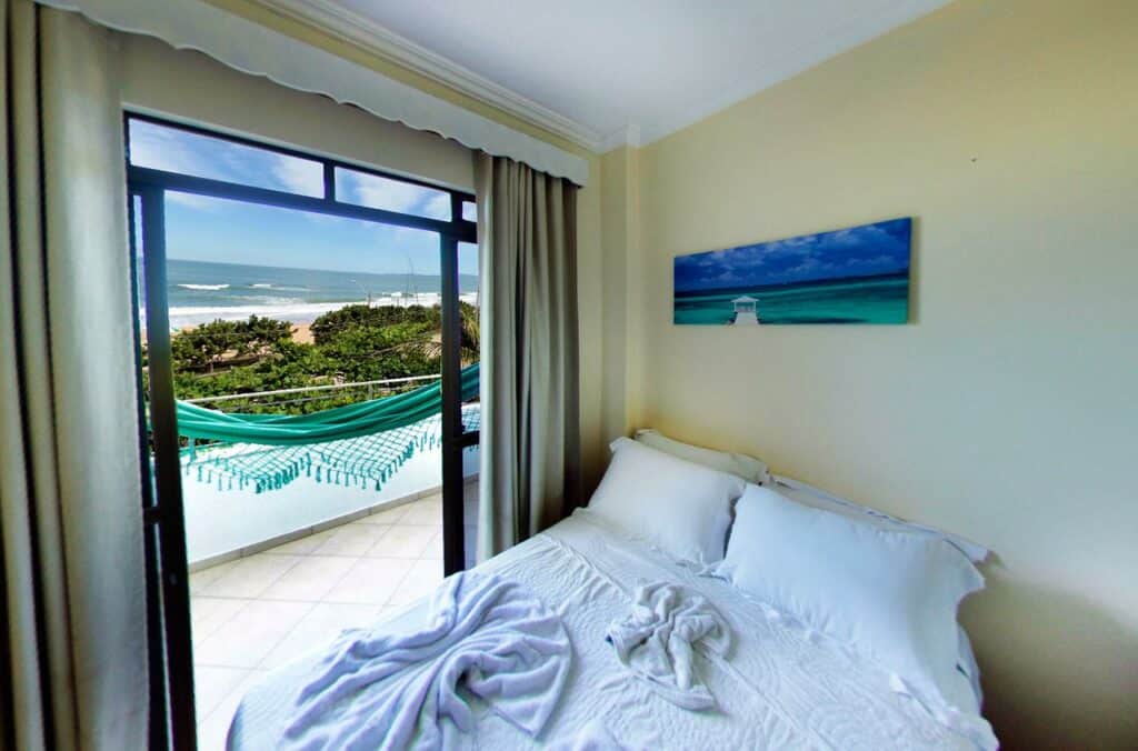 Quarto da Pousada OceAnas Frente Mar Estaleiro, com uma cama de casal ao lado de uma porta de vidro que dá acesso à sacada, que tem uma rede verde e vista para o mar