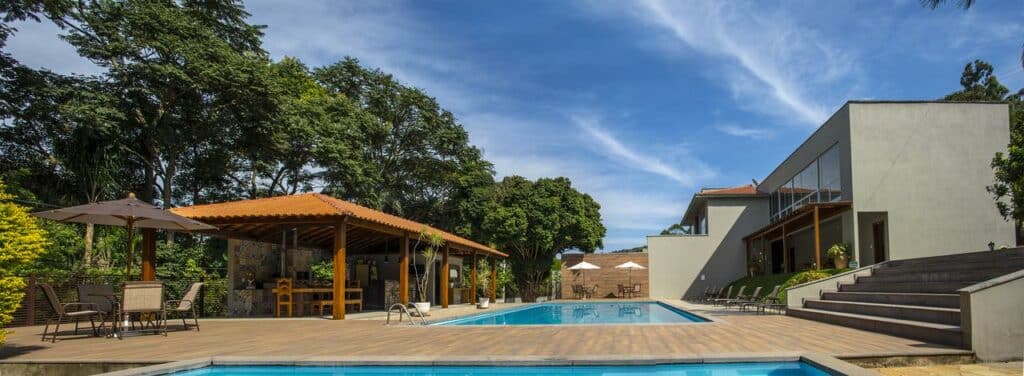 Área da piscina externa com espreguiçadeiras da Pousada Recanto das Araras, uma das pousadas em Tiradentes, assentos à sombra sob uma pérgola e um edifício moderno cercado por árvores.