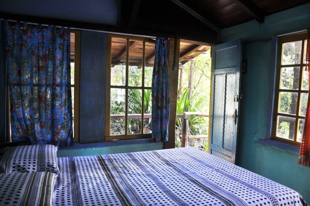 Quarto da Pousada Rural Lua Bonita do Nero, com uma cama de casal, janelas com cortinas azuis floridas e a porta aberta mostrando a natureza exuberante do lado de fora