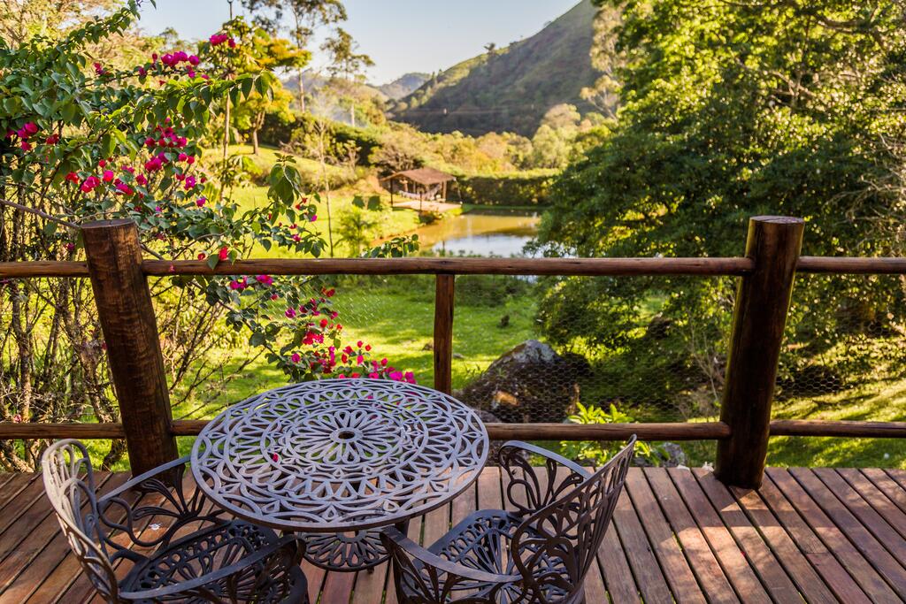 Varanda de uma das pousadas na Serra da Mantiqueira, com um deck de madeira, mesa redonda com duas cadeiras e uma vista para um lago marrom, com bastante gramado verde, árvores e flores em volta