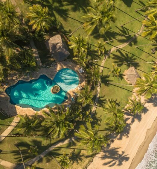 vista aérea da Pousada Xalés de Macaraípe, com uma piscina no meio de uma área verde com gramado e coqueiros. À direita tem uma praia