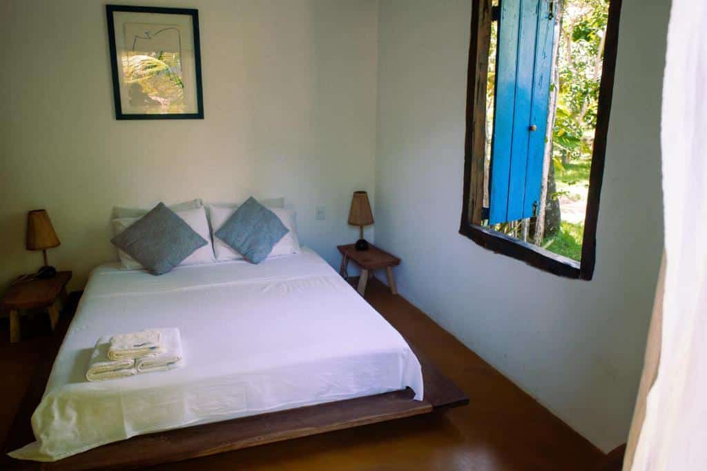 Quarto com cama de casal, lençóis brancos, almofadas azuis e janela também azul, no Sítio Peixe do Mato, uma das pousadas em São Miguel dos Milagres
