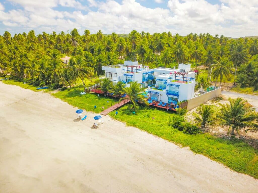 Edifício da Sollaris Ecopousada, com bastante natureza verde em volta e um corredor estilo deck de madeira dando acesso à areia branca da praia, para ilustrar as pousadas em Alagoas