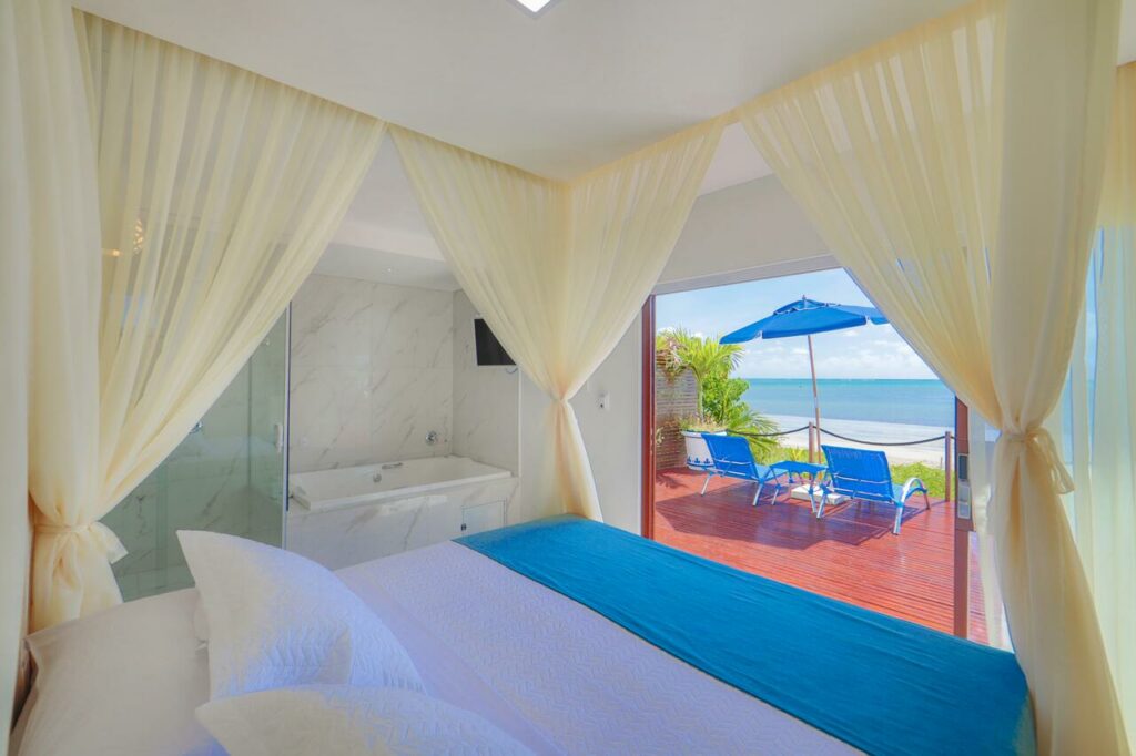 Quarto da Sollaris Ecopousada, com cama de casal, hidro e uma varanda privativa com duas espreguiçadeiras, um guarda-sol e vista para a praia de água azul
