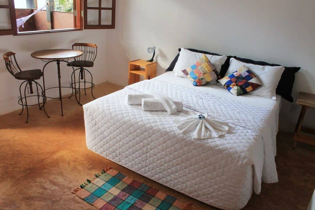 Quarto da Tiô Isolda Artes & Hospedaria, com uma cama de casal com duas toalhas, travesseiros e almofadas coloridas em cima. No lado direito há uma mesa redonda com dois bancos e uma janela mostrando a varanda com rede
