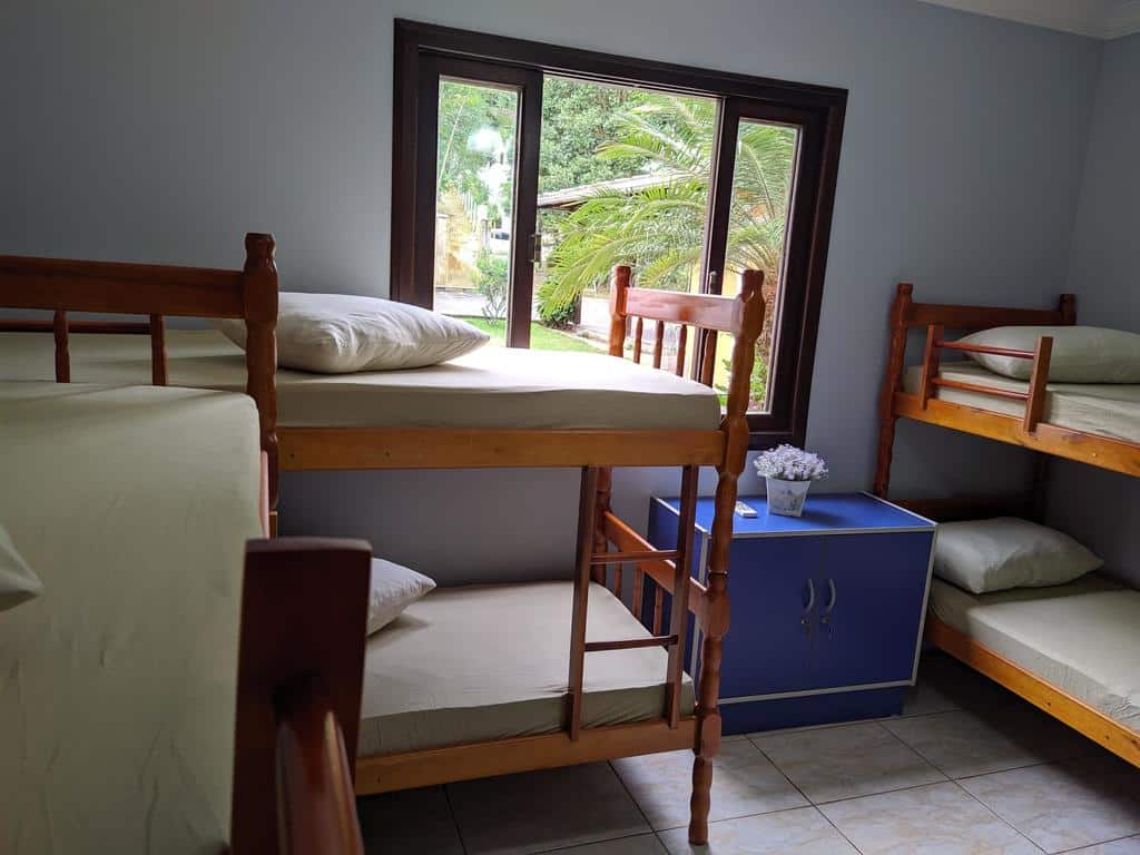 Interior de um quarto misto da iUP HOUSE Hostel, com três camas de beliche, um armário pequeno azul com um vaso de flor em cima e uma janela mostrando o jardim no lado de fora
