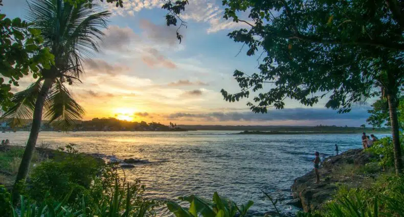 Pôr do sol em um rio com árvores emoldurando a vista, pessoas em pé na margem rochosa e nuvens no céu, na Vila do Dengo em Itacaré.