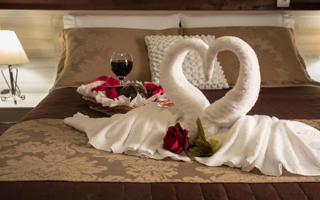 cama de casal com uma escultura de dois cisnes feita em toalhas brancas em cima da cama. Há também duas taças de vidro e uma cesta com pétalas de rosas.