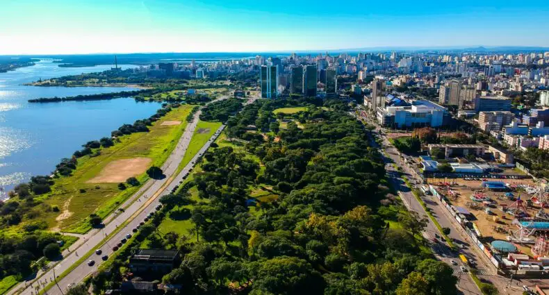Vista aérea de uma cidade com um rio, um parque verde, edifícios altos e uma estrada movimentada que passa ao lado do parque. Representa as pousadas em Porto Alegre