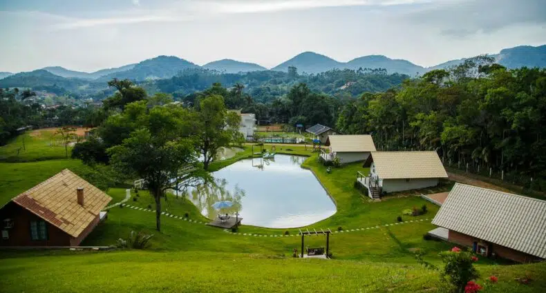 Várias casas pequenas com telhados inclinados cercam um lago, situado em uma paisagem gramada com colinas e árvores ao fundo.