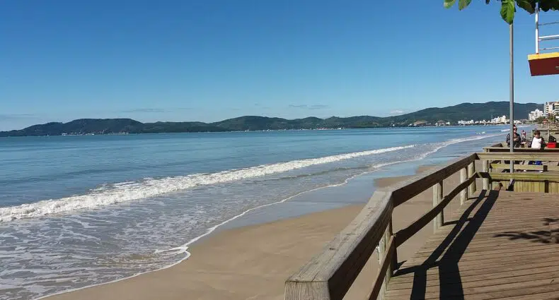 Um calçadão de madeira corre paralelo a Média Praia com areia com ondas suaves, sob um céu azul claro e colinas distantes, para ilustrar o post de pousadas em Itapema.