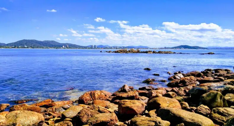 Praia da Armação com um litoral rochoso com mar azul claro, montanhas distantes e nuvens brancas espalhadas sob um céu azul brilhante. Ilustra o post de pousadas em Penha SC