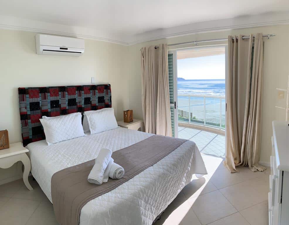 Quarto da Pousada Azores, de 15 m², com uma cama, uma escrivaninha branca de cada lado da cama, e uma porta dando acesso à sacada do quarto com vista do mar