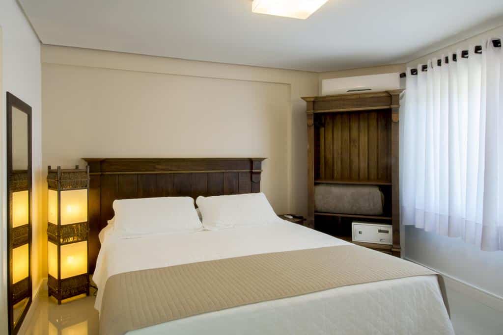 Interior do Quarto Deluxe da Pousada Bora Bora, de 35 m², com uma cama, uma iluminaria artesanal ao lado, um espelho e um armário de madeira com um cofre branco e um ar-condicionado a cima