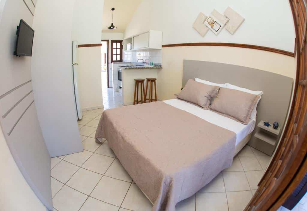 Um pequeno quarto da Pousada Brisa do Mar com uma cama de casal, TV na parede e uma cozinha compacta com dois bancos de bar ao fundo.