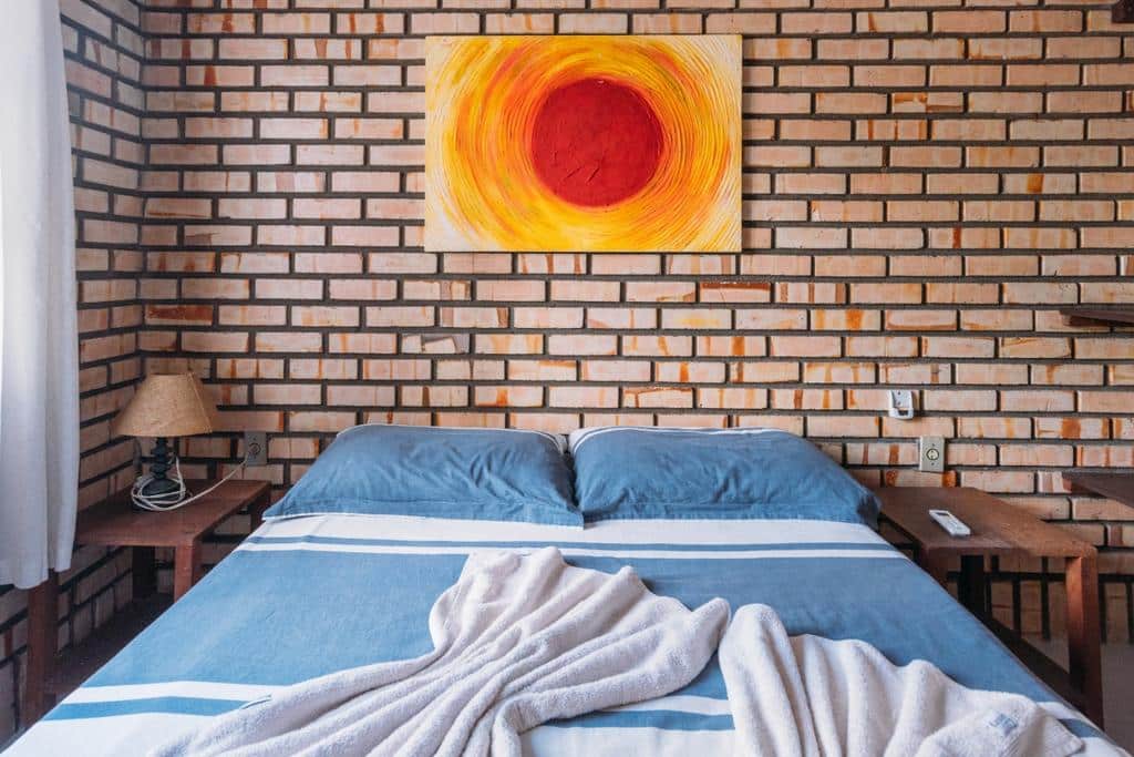 Quarto da Pousada Canto das Trilhas, com parede de tijolinhos, um quadro de espiral degradê amarelo e vermelho decorando o espaço, e uma cama com colcha azul, dois travesseiros e duas toalhas em cima