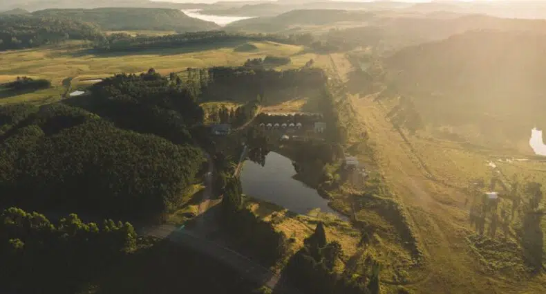 Vista aérea de uma paisagem rural na Pousada Recanto do Lago com colinas, campos, um pequeno lago e edifícios espalhados sob um céu nebuloso. Ilustra o post de pousadas em Cambará do Sul.