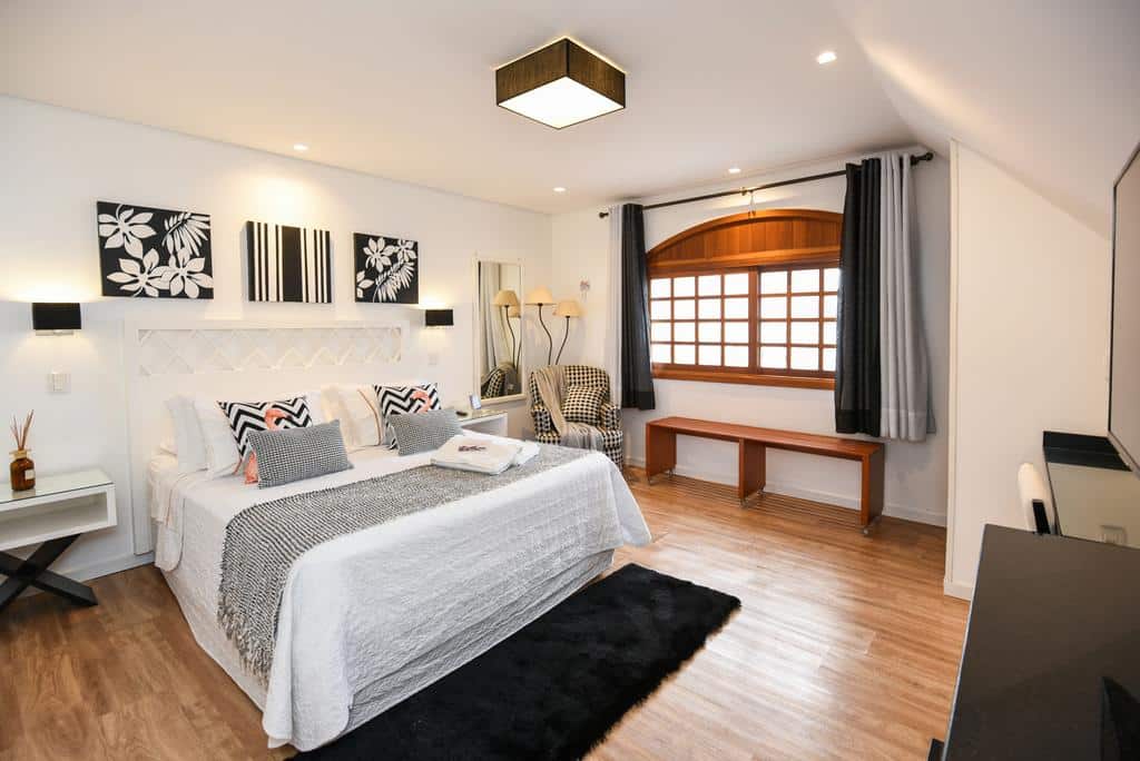 quarto da Pousada La Lavande com uma cama de casal à esquerda, uma poltrona xadrez preto e branco no cano e uma janela ampla de madeira na parede ao fundo