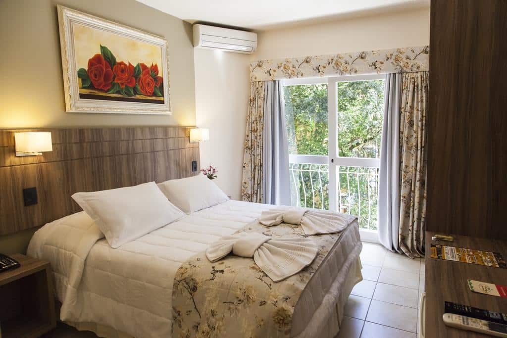 suíte da Pousada Águia Branca com uma cama de casal à esquerda, um quadro de rosas pendurado acima da cabeceira e portas duplas com acesso a sacada privativa e vista para o jardim