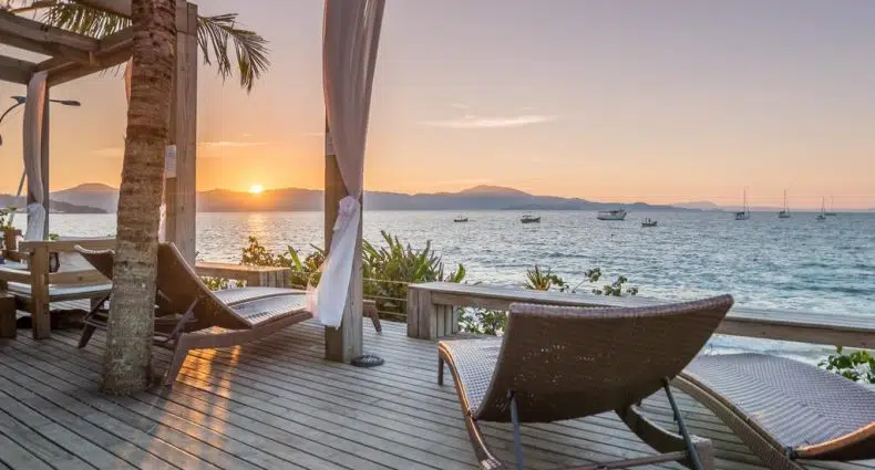 Deck de madeira da Pousada dos Sonhos, uma das pousadas em Jurerê Internacional com espreguiçadeiras com vista para o oceano ao pôr do sol, com veleiros na água e montanhas à distância.